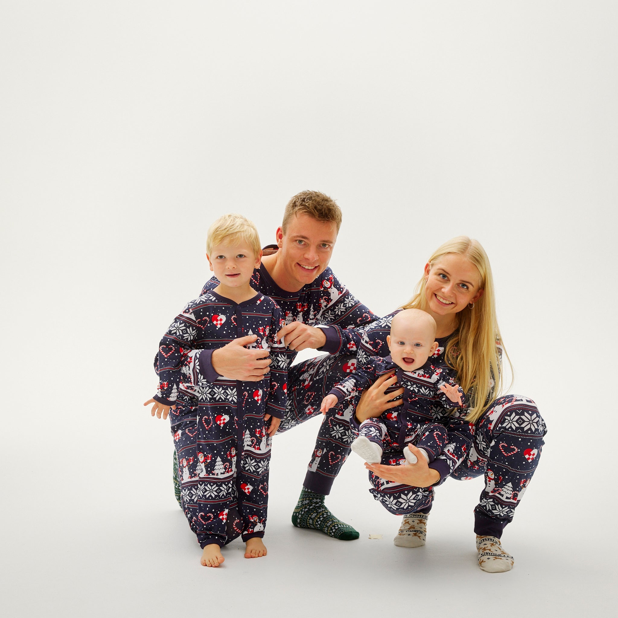 Drei Personen in passenden dunkelblauen Weihnachtspyjamas mit Herz- und Schneeflockenmuster, Baby, Mann und Frau lachend.